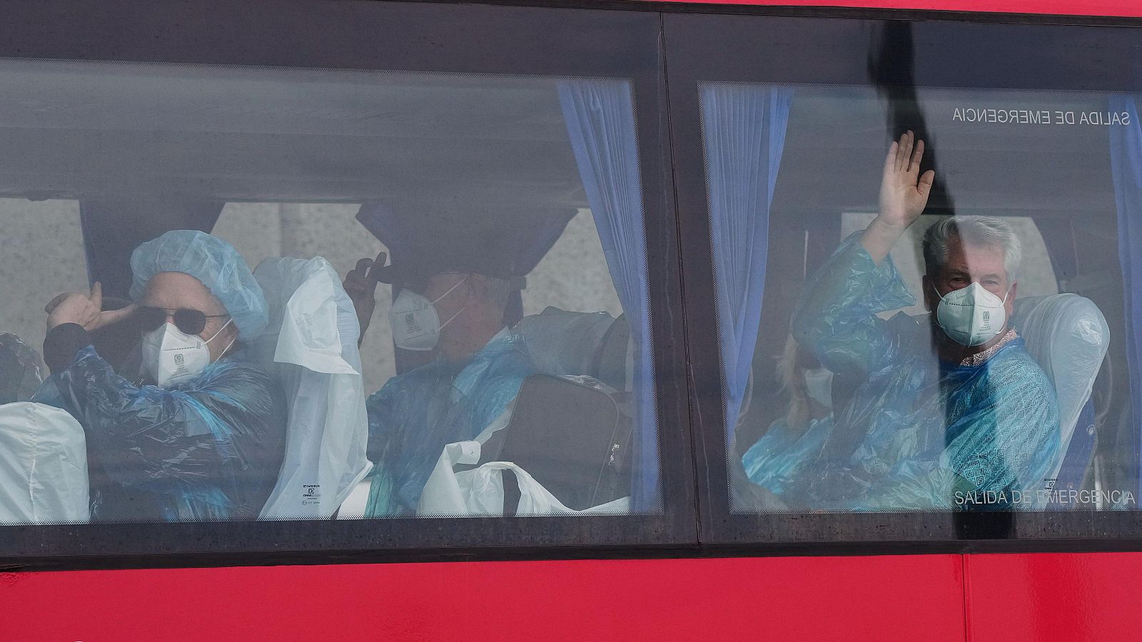Pasajeros con mascarillas y batas azules saludan desde un autobús rojo. Se observa "SALIDA DE EMERGENCIA" invertido en la ventana.