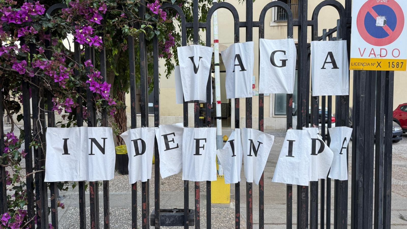 Verja negra con carteles de "VAGA INDEFINIDA" en el IES Llúis Vives, adornada con vegetación púrpura y señales de tráfico.