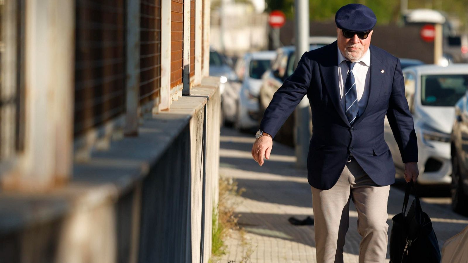 José Villarejo, con barba blanca, gafas de sol y traje azul marino, camina por una acera urbana con un bolso oscuro. Se distingue una señal de tráfico al fondo.