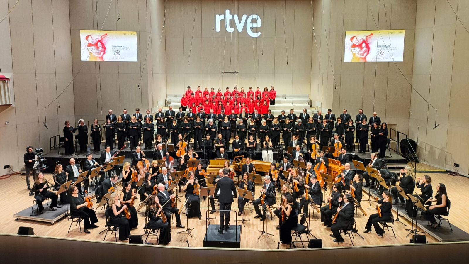 Gran escenario con la Orquesta y Coro RTVE actuando. Coro en gradas (rojo/negro), orquesta en frente (negro). Fondo con logo RTVE y pantallas: "Temporada 2023-2024".