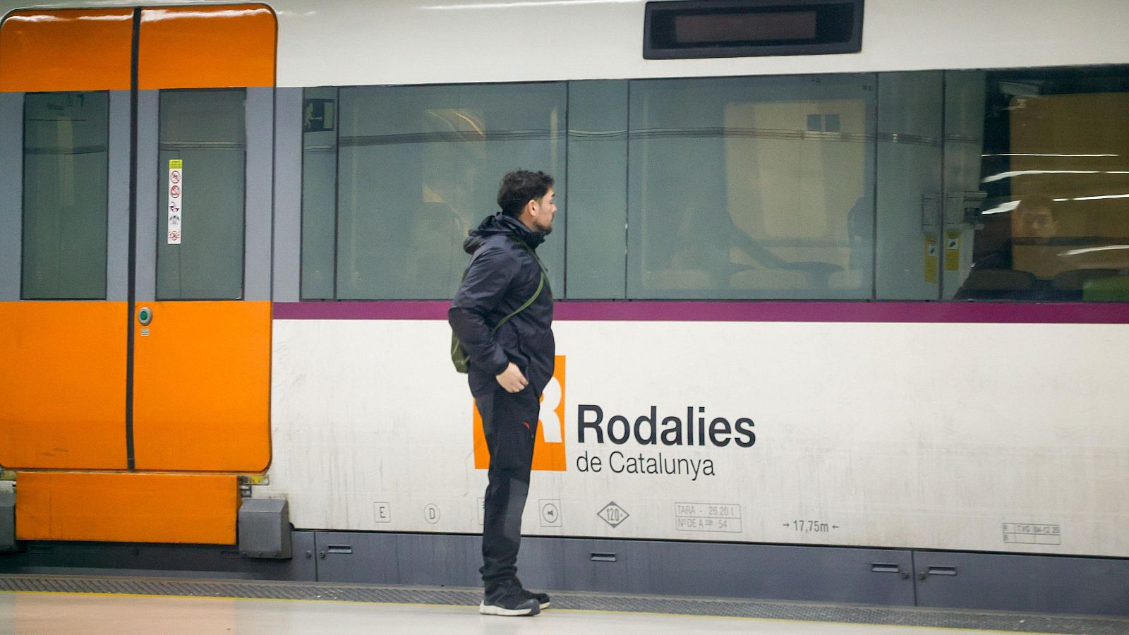 Home dempeus a l'andén al costat d'un tren de Rodalies de Catalunya, amb jaqueta fosca, motxilla i portes taronges.