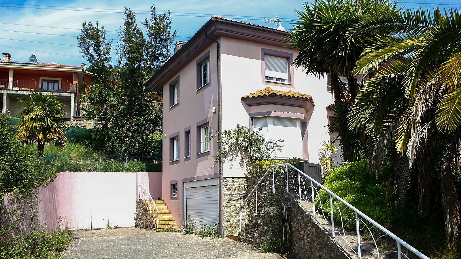 Casa de tres pisos, rosa y blanco, con puerta de garaje blanca y escalera exterior. Vegetación y muro rosa rodean la propiedad.