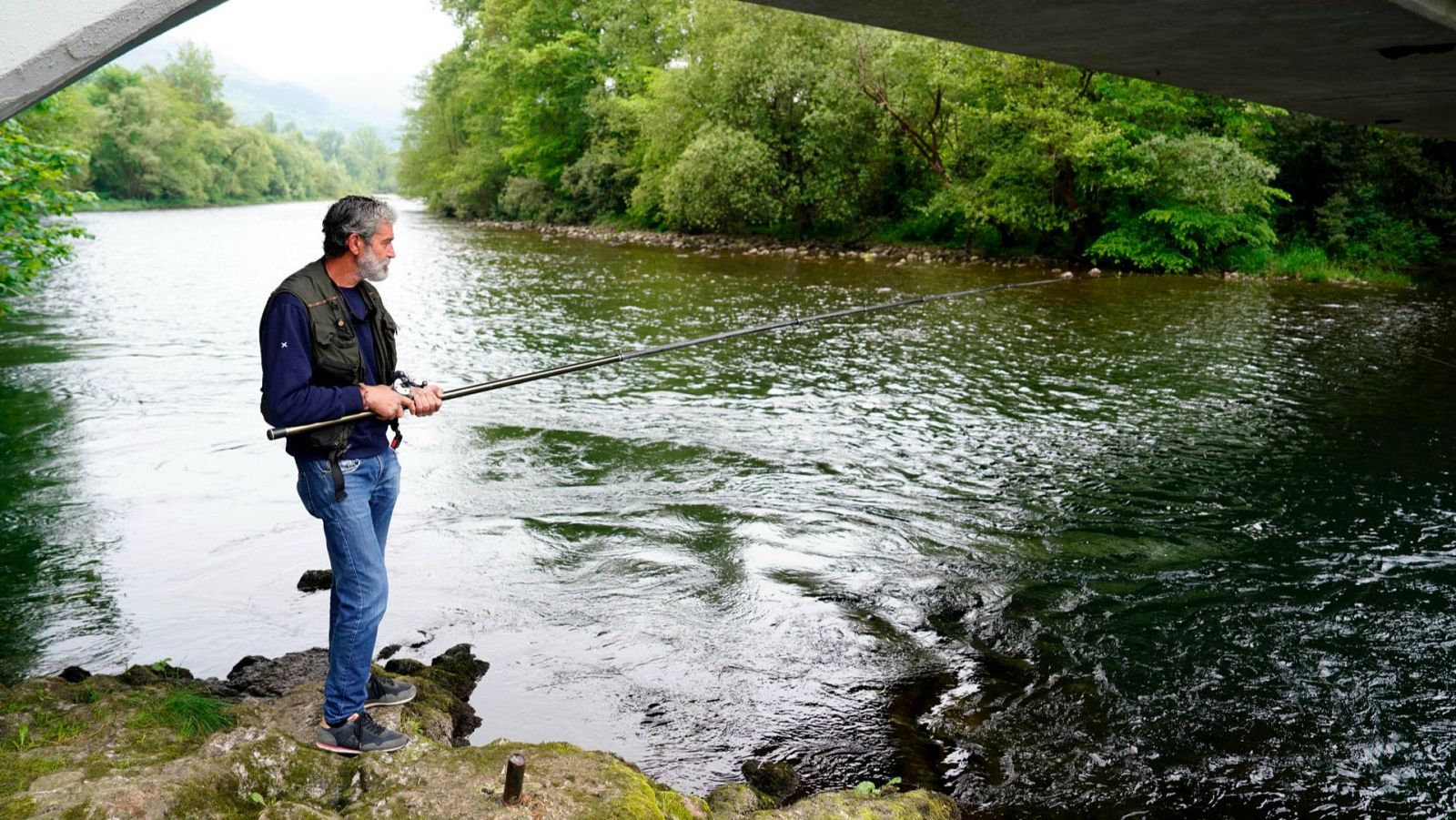 Pescador con barba canosa, vestido con chaleco de pesca, espera pacientemente en la orilla de un río con su caña.