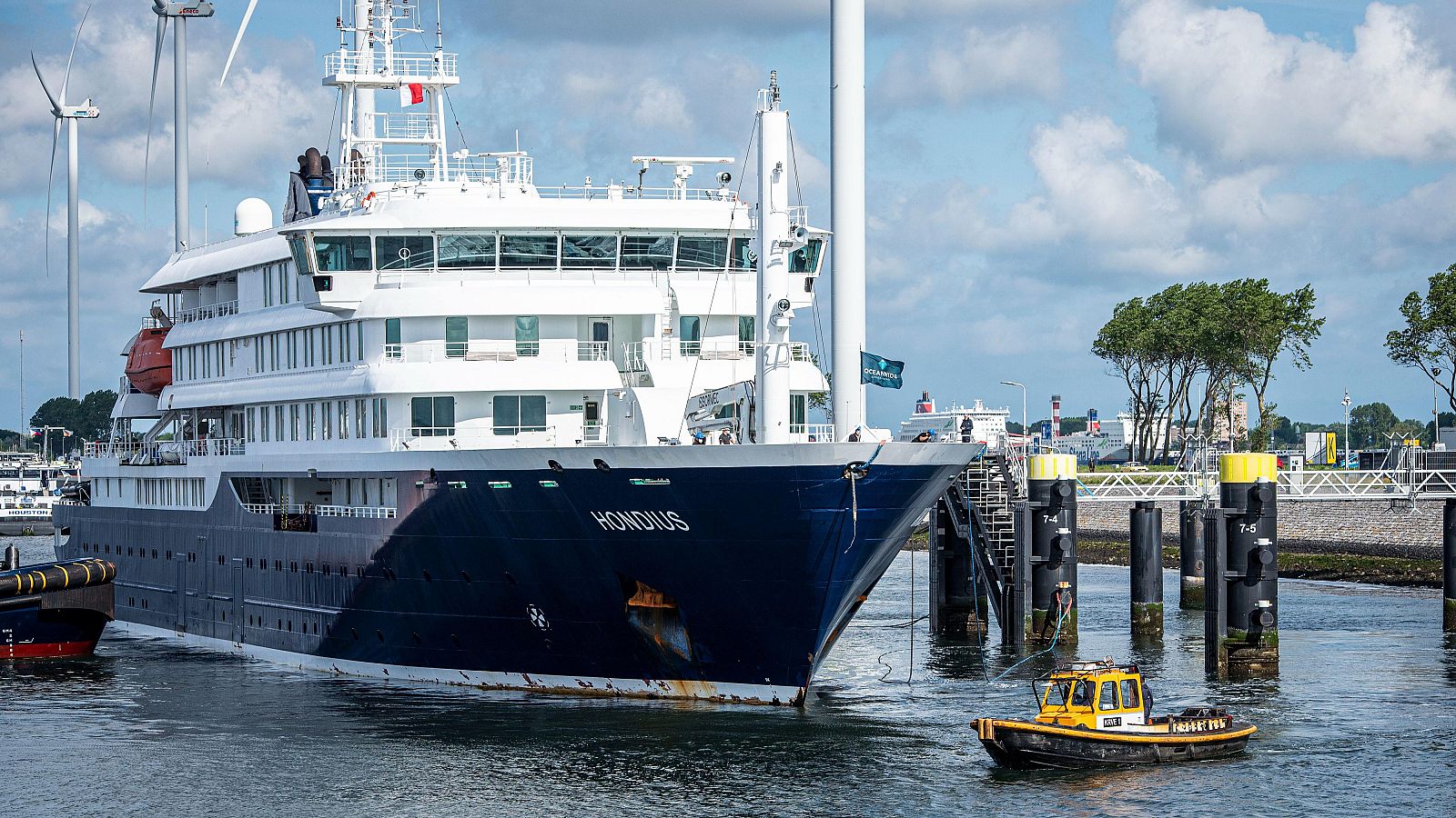 El M.V. Hondius atraca en puerto, con su nombre visible en la proa, botes naranjas y turbinas eólicas al fondo.