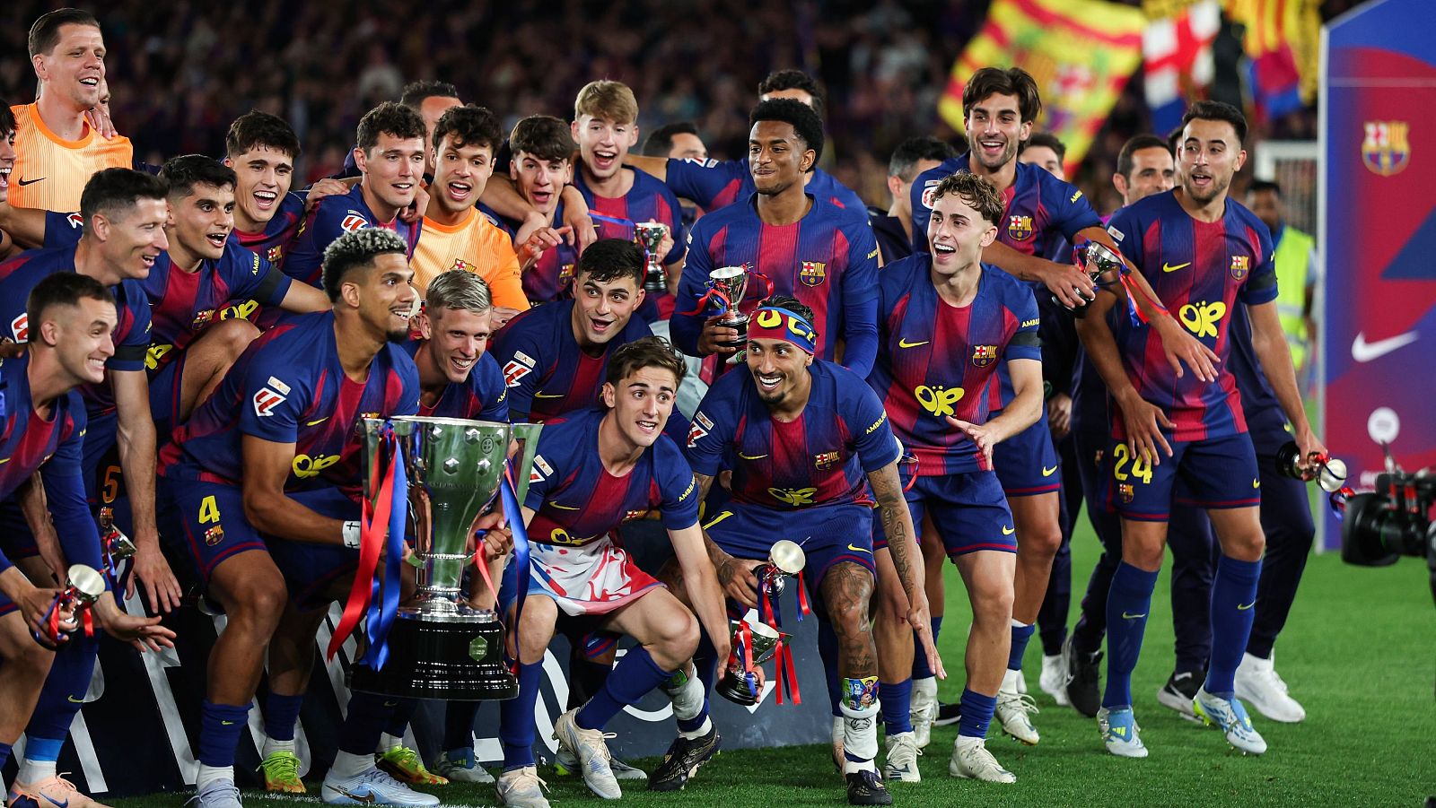 Jugadores del FC Barcelona celebran un título de liga con el trofeo. Se ven Jules Kounde, Ronald Araújo, Gavi, Pedri y Raphinha entre otros, todos sonriendo y mostrando euforia en un campo de césped.