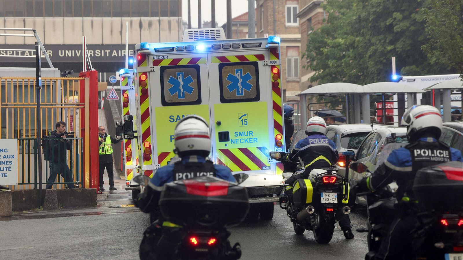 Ambulancia del SMUR NECKER con luces azules, escoltada por policías en moto, se dirige a un hospital universitario. El suelo está mojado.