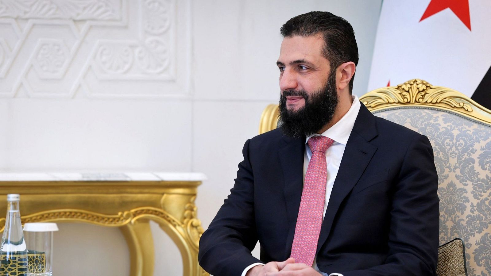 Ahmed al-Sharaa, presidente sirio, en traje oscuro y corbata rosa, sentado en silla clásica con mesa auxiliar y bandera de Siria al fondo.