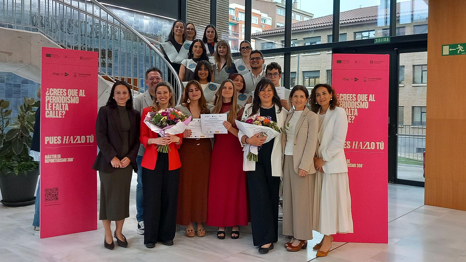 Graduación del Máster de Reporterismo 360º: grupo de graduados con estolas, ramos y diplomas, junto a un cartel rosa con el lema "¿CREES QUE AL PERIODISMO LE FALTA CALLE? PUES HAZLO TÚ".