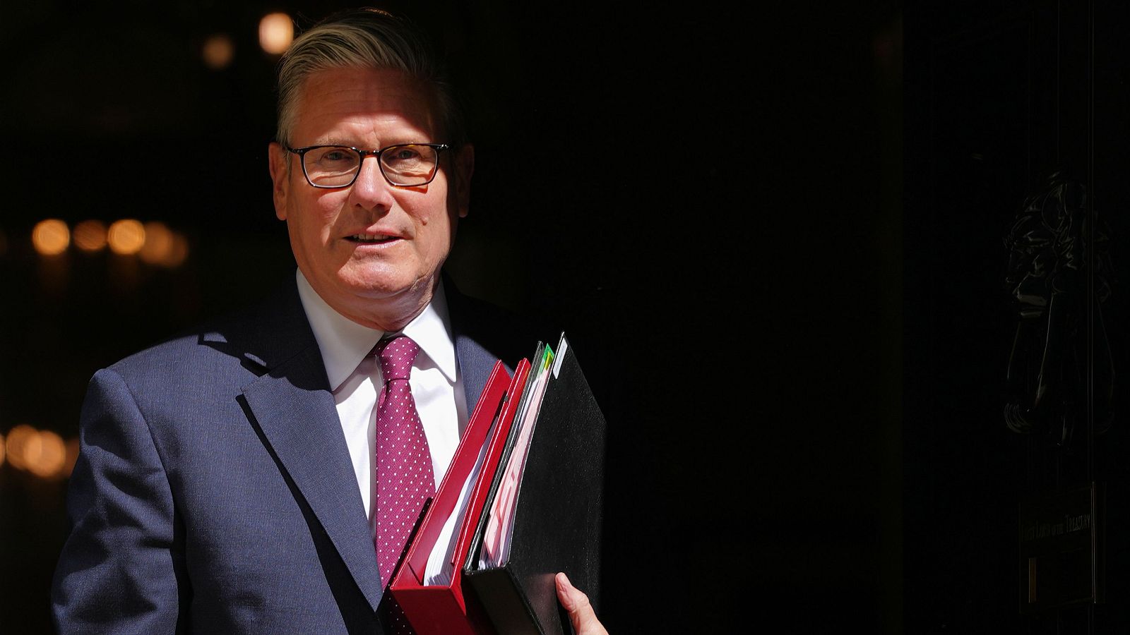 Primer plano de Keir Starmer, líder laborista británico, con traje azul marino, camisa blanca y corbata granate, sosteniendo carpetas. Mira al frente con expresión seria.