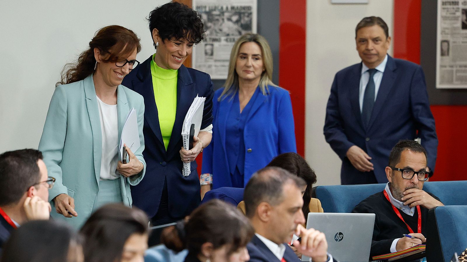 Elma Saiz y Mónica García, ministras, posan en una sala institucional con otras personas. Se aprecian detalles de sus atuendos y expresiones faciales.