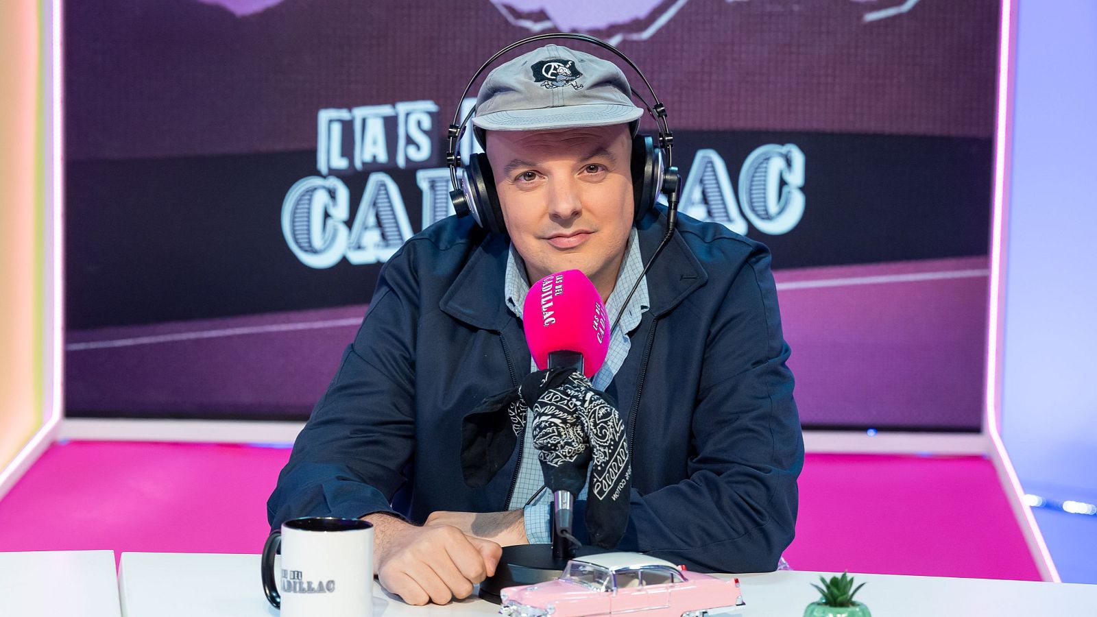 Carlangas en un estudio de radio con micrófono rosa, taza y pantalla digital con "LAS DEL CADILLAC".