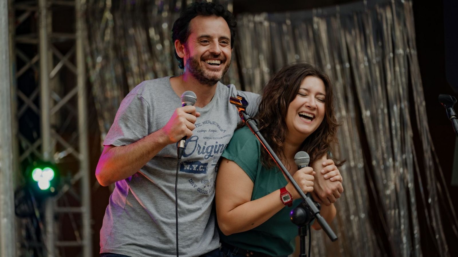 Dani Rovira y Clara Lago ríen juntos en un evento, ambos con micrófonos. Él lleva camiseta gris 'N.Y.C. Original', ella camiseta verde y reloj rojo.