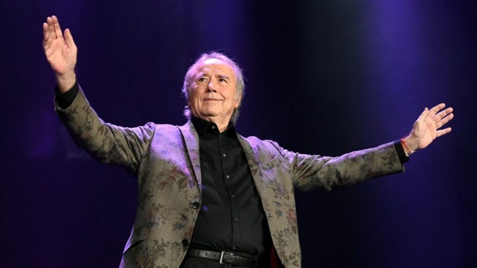 Joan Manuel Serrat, con cabello canoso, viste camisa negra y blazer estampado. Sus brazos están extendidos en un gesto de saludo o agradecimiento, con una leve sonrisa. Fondo oscuro con luces púrpuras y azules.