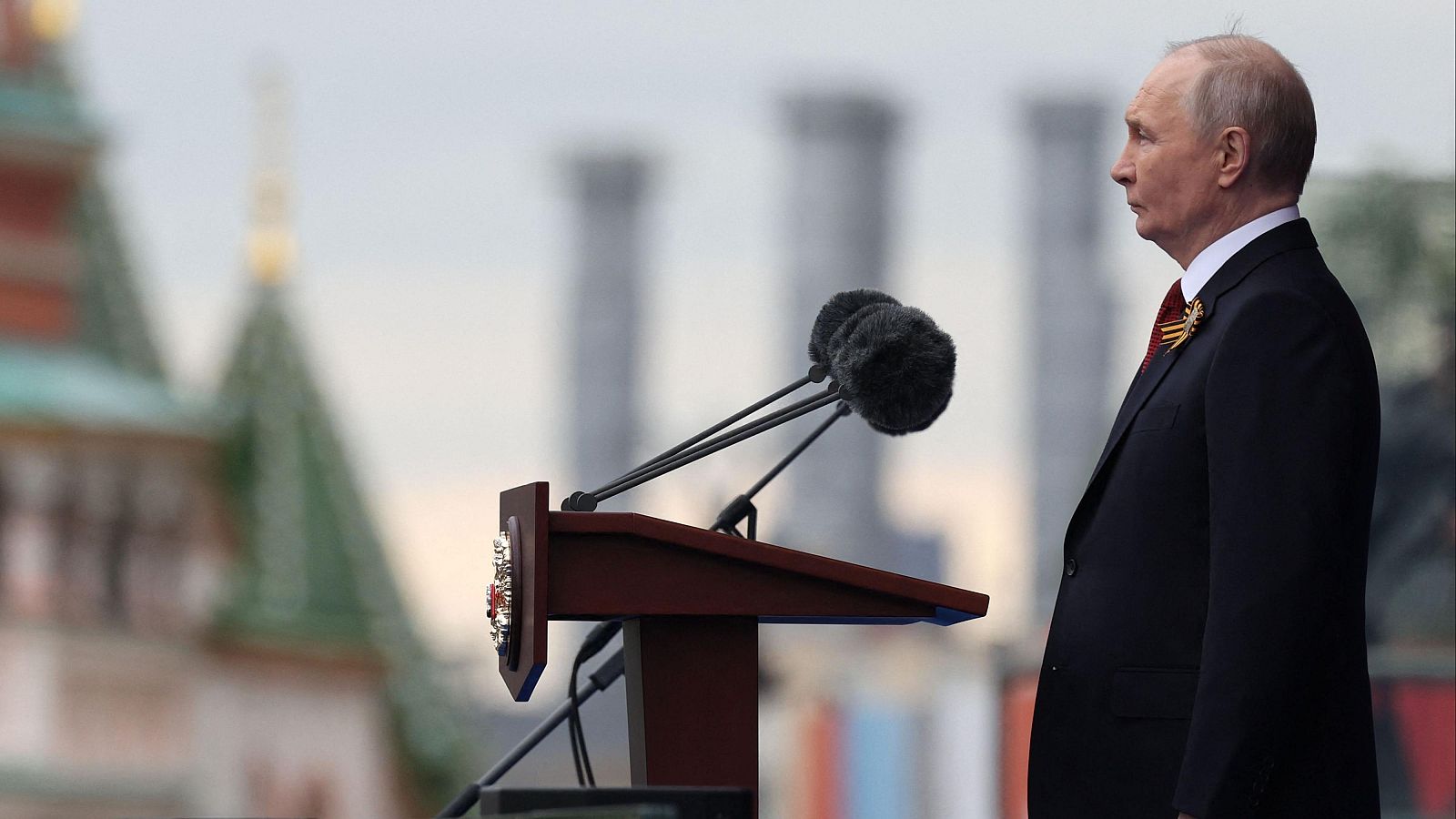 Vladímir Putin, de perfil, con traje oscuro y condecoración, junto a un atril con micrófonos. Fondo con arquitectura tipo Kremlin y chimeneas industriales.