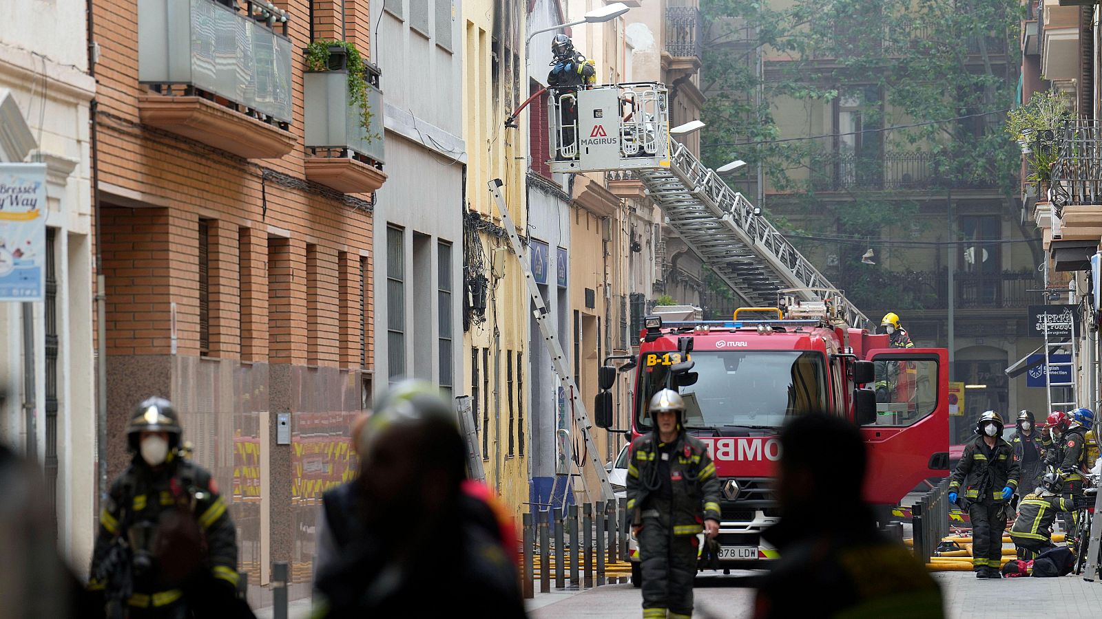 Bomberos con equipo de protección actúan en una calle urbana tras un incendio en un edificio, con un camión MAGIRUS y su escalera desplegada.