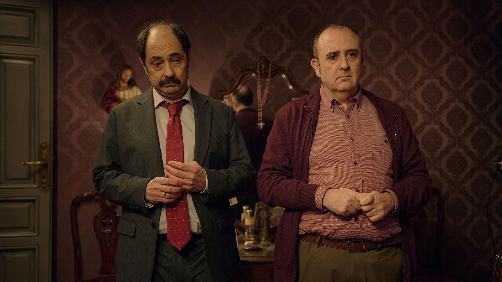 Jordi Sánchez y Carlos Areces, actores españoles, posan en una habitación decorada. Sánchez viste traje gris y corbata roja; Areces, cárdigan burdeos y camisa rosa.