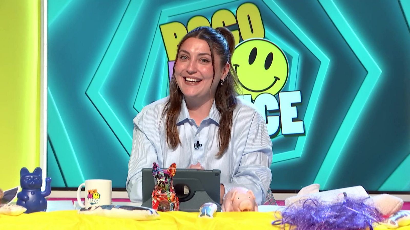 Marina Lobo sonríe en un estudio de TV con fondo turquesa y neón azul. El logo "POCO ME PARECE" con un smiley es visible. En primer plano, una mesa con objetos coloridos y una tablet.