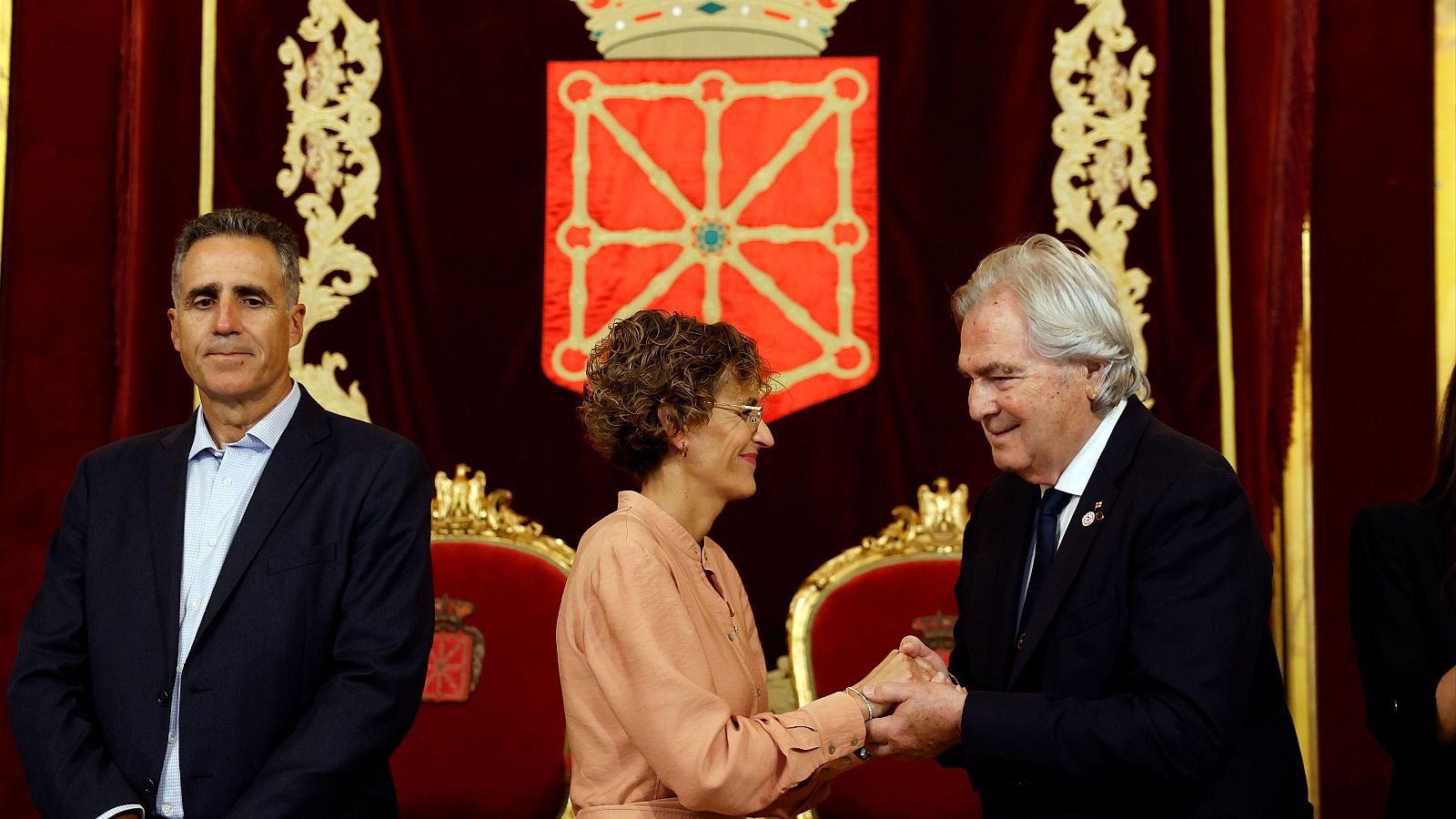 María Chivite, presidenta de Navarra, saluda a Gian Francesco Lupatelli, presidente de ACES Europe, con Miguel Induráin presente. Fondo: tapiz con escudo de Navarra.