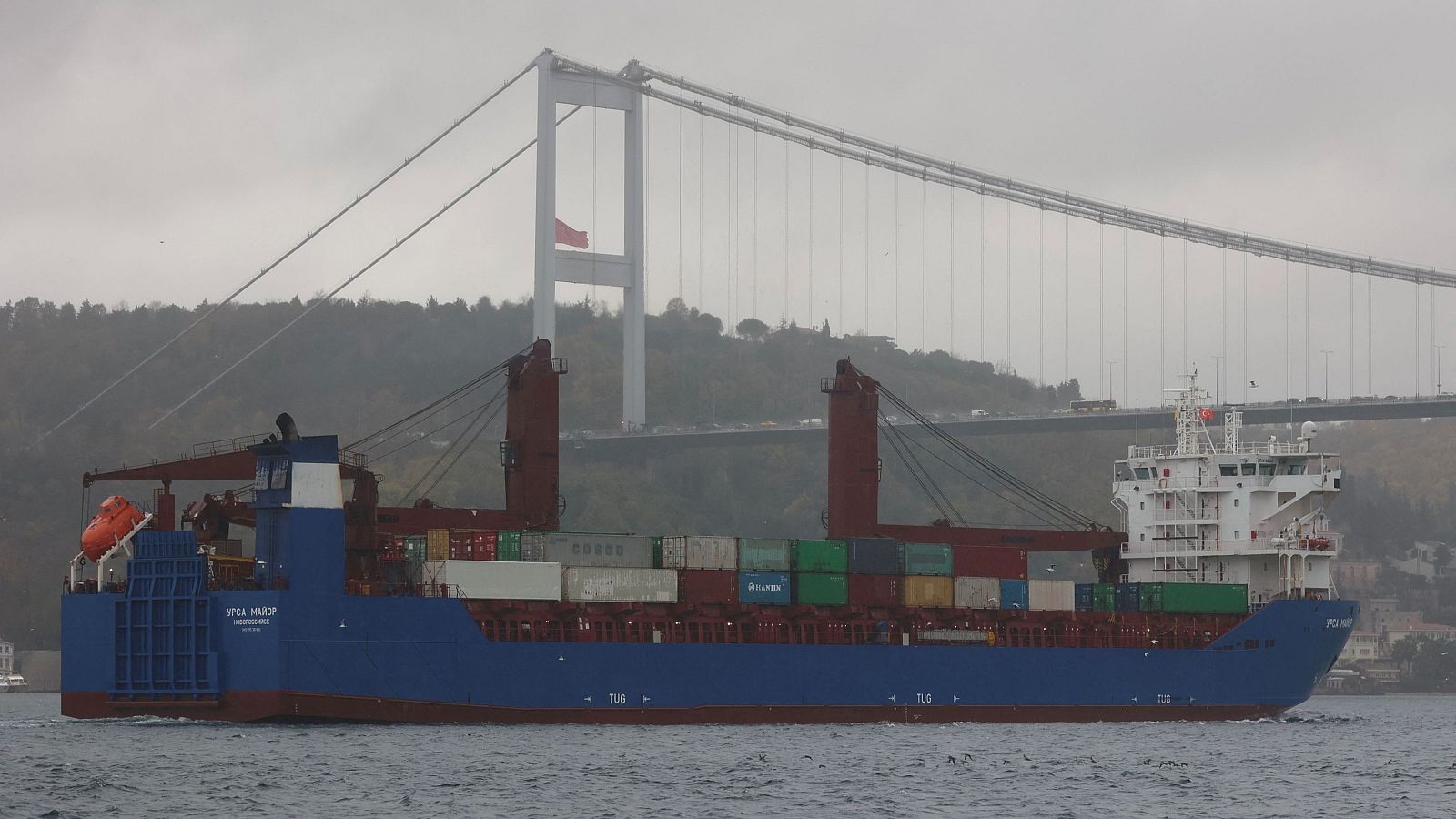 Carguero Ursa Major navegando, casco azul oscuro y blanco, transportando contenedores. Cielo nublado.