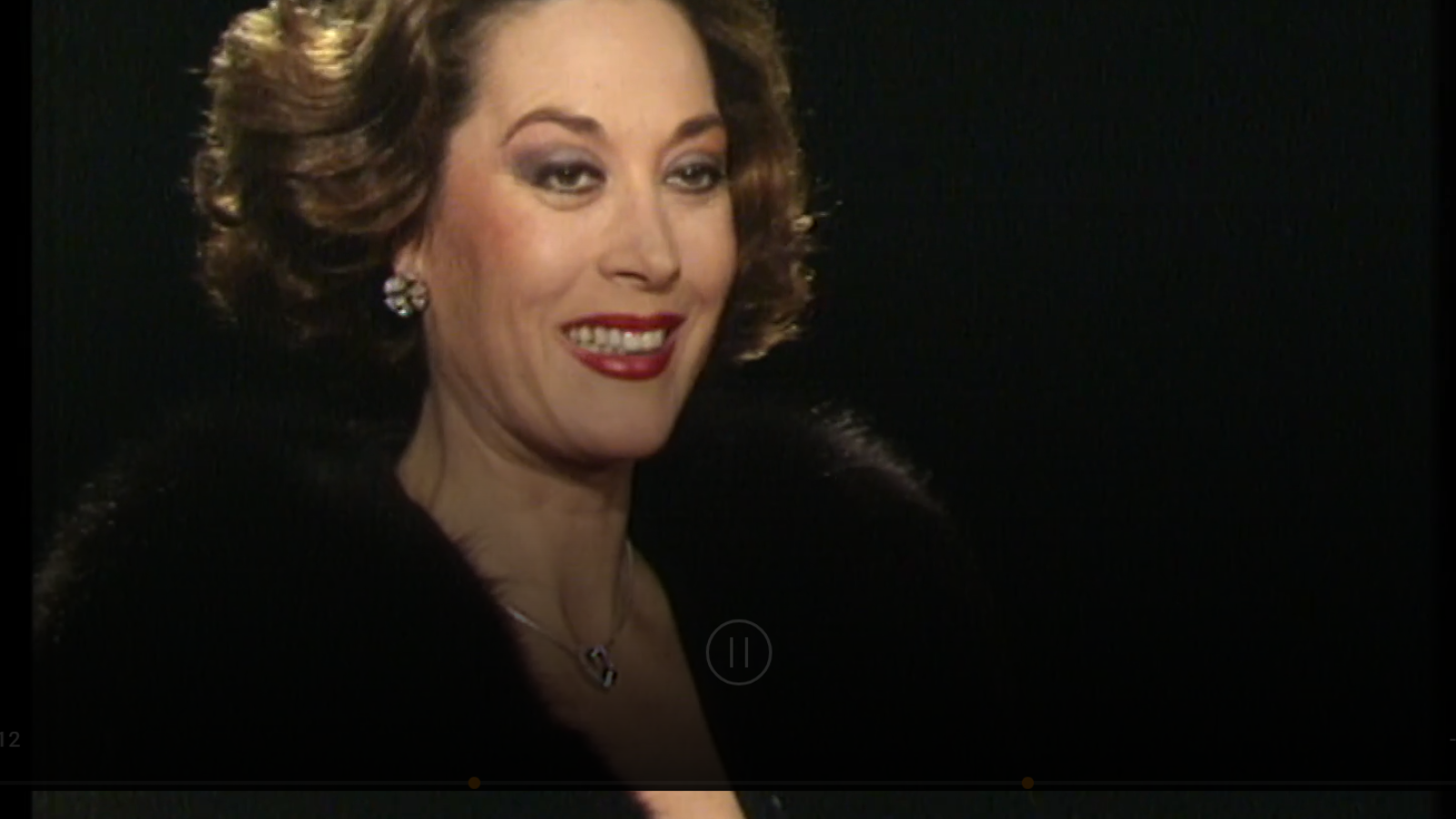 Tania Doris sonríe en un retrato de archivo de RTVE. Cultura, con peinado voluminoso, maquillaje y joyas. Fondo negro con elementos de reproductor de vídeo.
