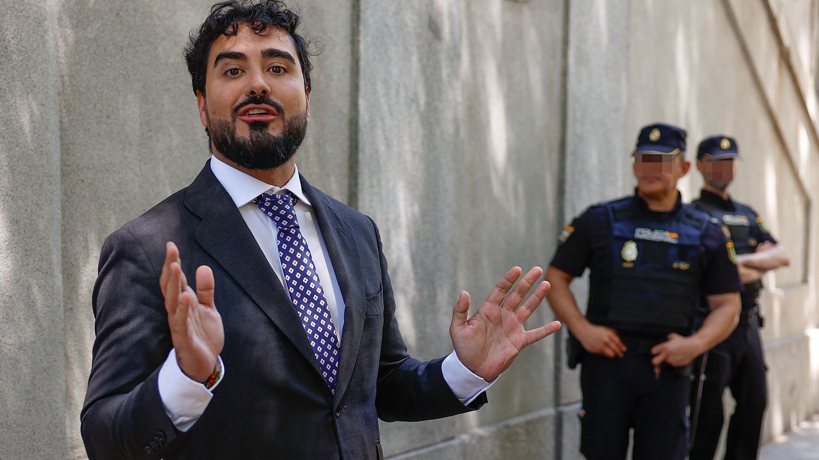 Alvise Pérez, vestido con traje y corbata, gesticula frente a un muro de piedra, con dos agentes de policía uniformados detrás.