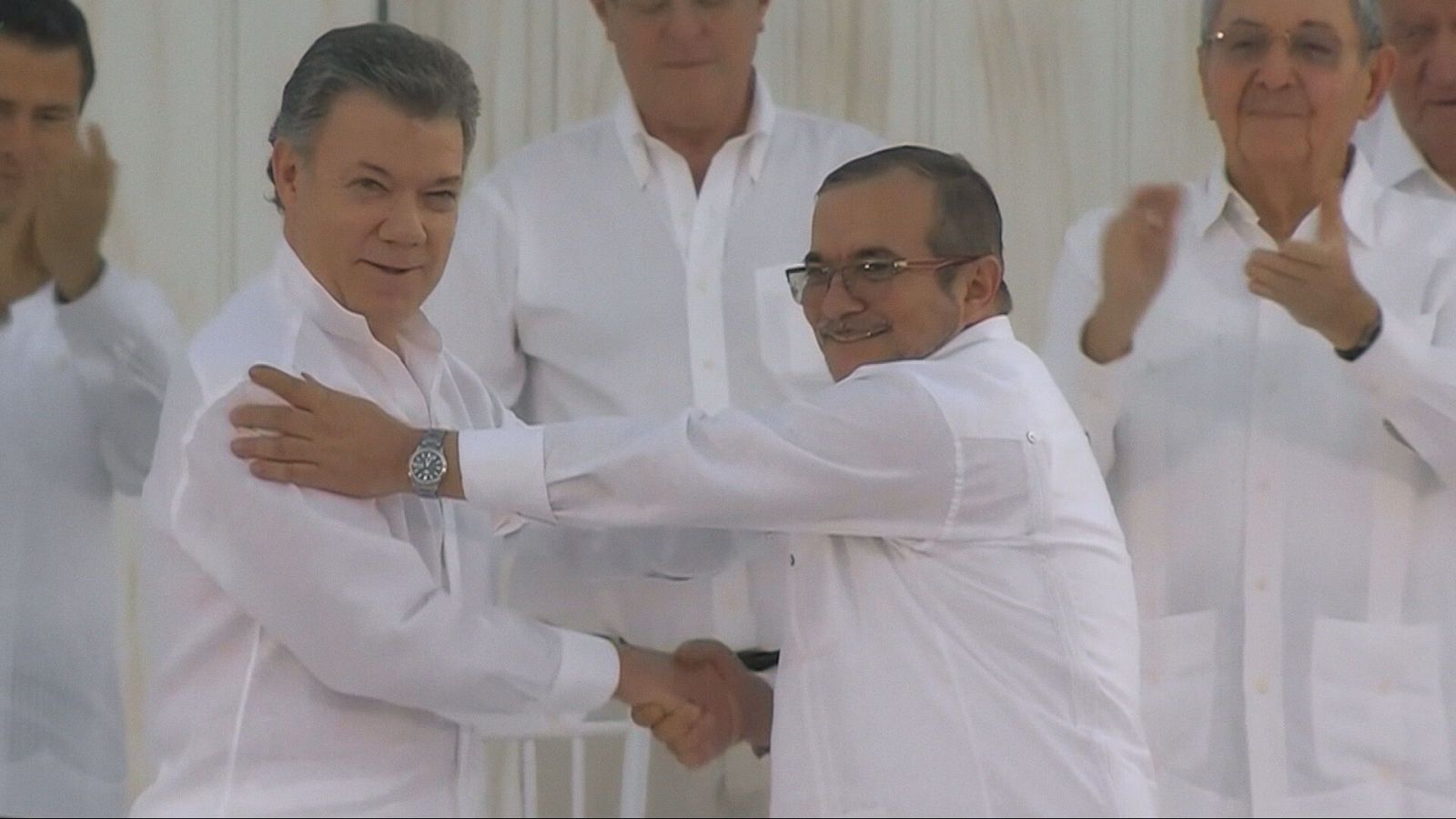 Juan Manuel Santos y Rodrigo Londoño, líderes de Colombia y las FARC respectivamente, sellan un acuerdo de paz en 2016. Ambos sonríen mientras se dan la mano, rodeados de asistentes que celebran el momento.
