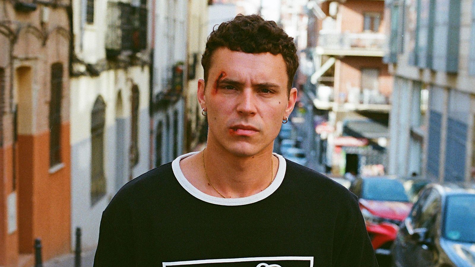 Aron Piper en primer plano, con heridas en el rostro, pendientes, collar y camiseta negra. Fondo de calle urbana con edificios antiguos.