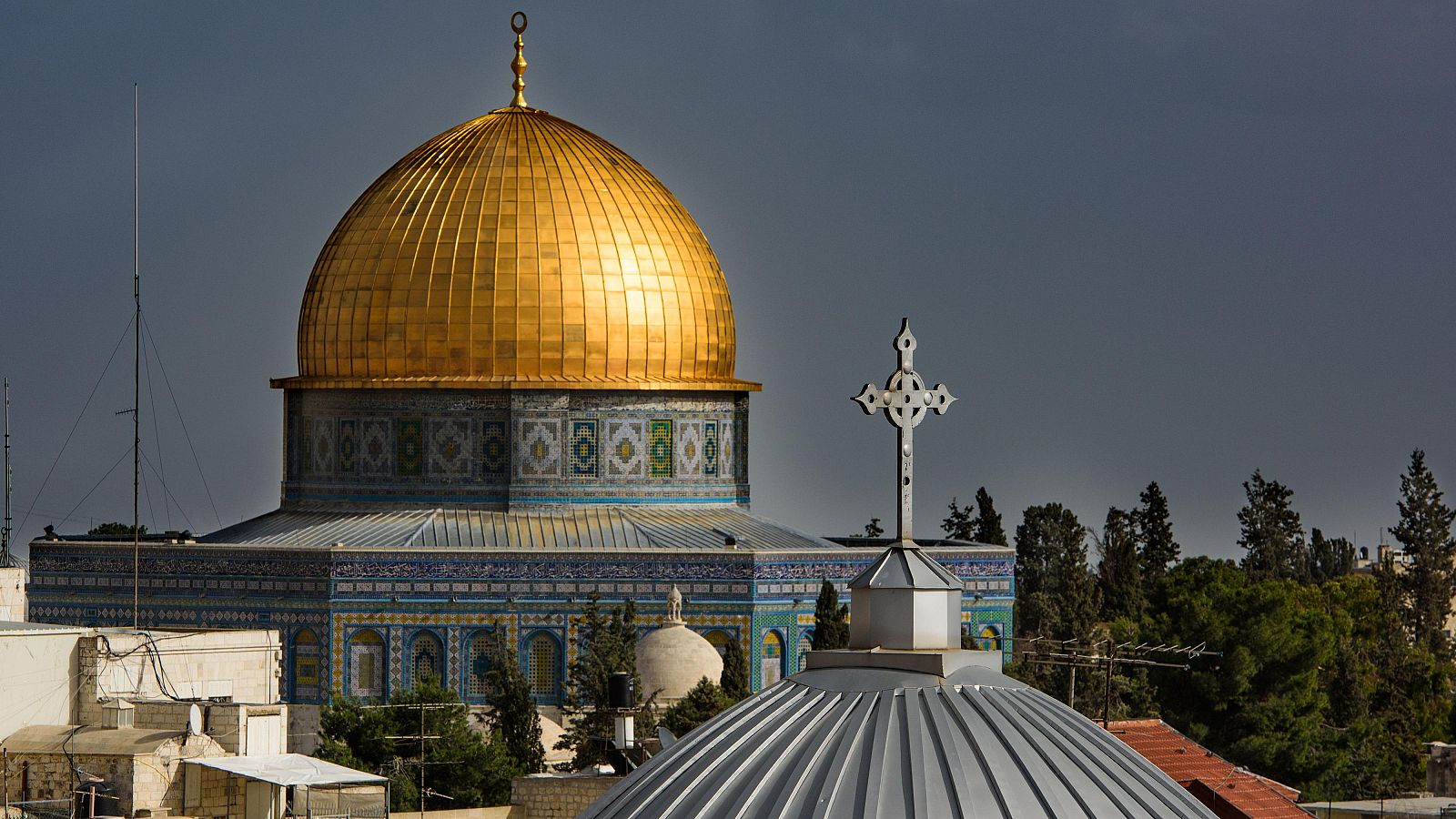 Vista panorámica de la Cúpula de la Roca y la Iglesia del Santo Sepulcro en Jerusalén, bajo un cielo nublado. Se aprecian detalles arquitectónicos y el entorno urbano.