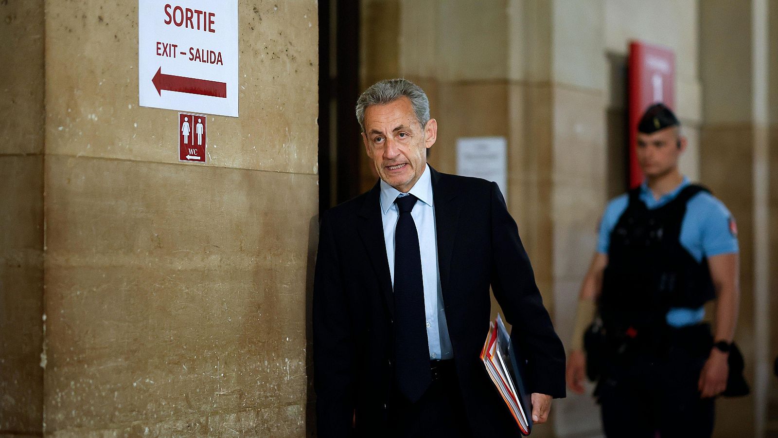 Nicolas Sarkozy, con traje oscuro y carpetas, se dirige por un tribunal en París, acompañado por un agente de seguridad. Señalización de salida y WC visible.