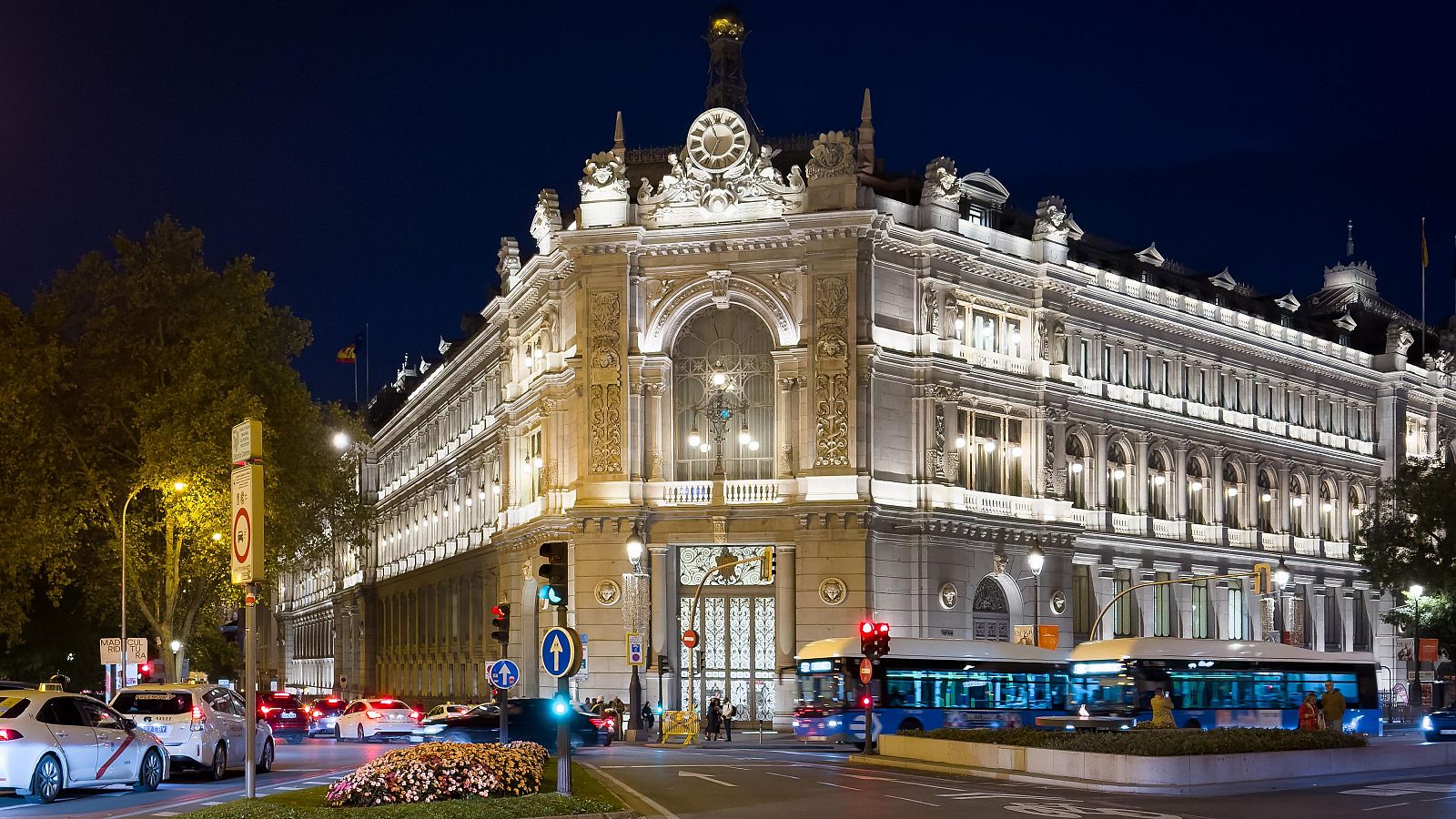 Vista nocturna de la Sede del Banco de España en Madrid, con tráfico vehicular y vegetación urbana iluminada.