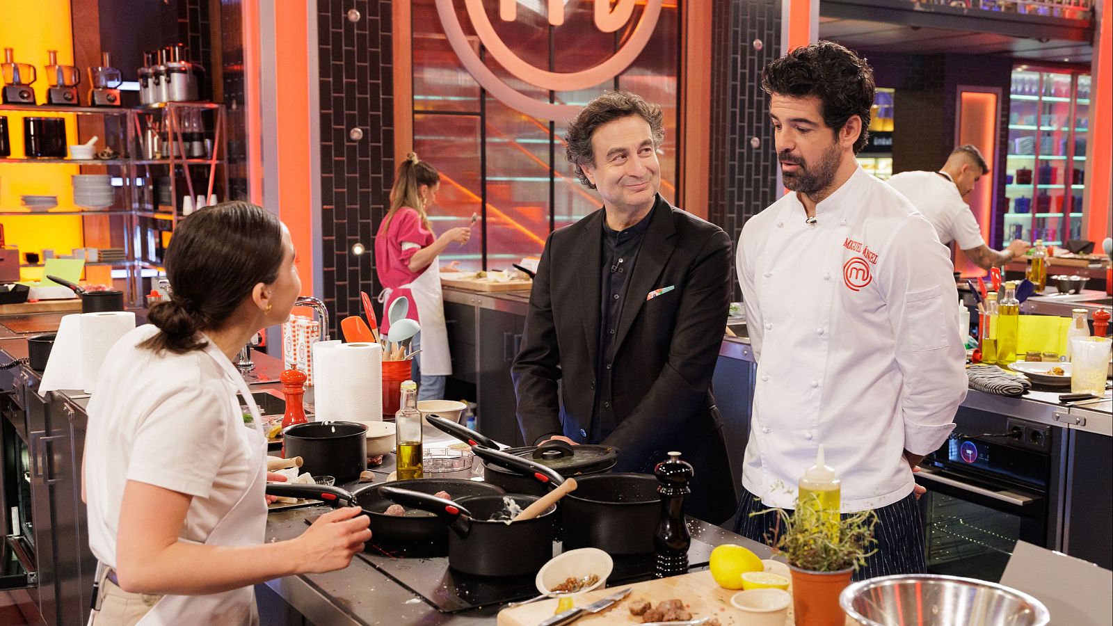 Miguel Ángel Muñoz y Jordi Cruz en el plató de MasterChef, interactuando con una concursante. Se aprecian detalles de la cocina profesional y otros participantes.