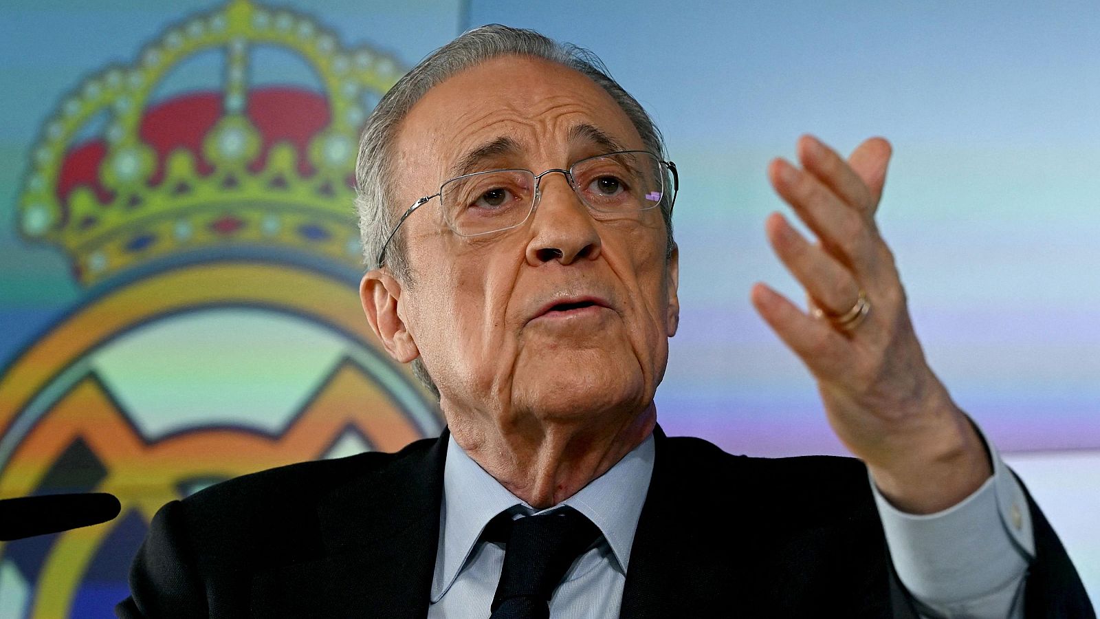 Primer plano de Florentino Pérez, presidente del Real Madrid, con traje oscuro y gesto explicativo. El escudo del club se ve al fondo.