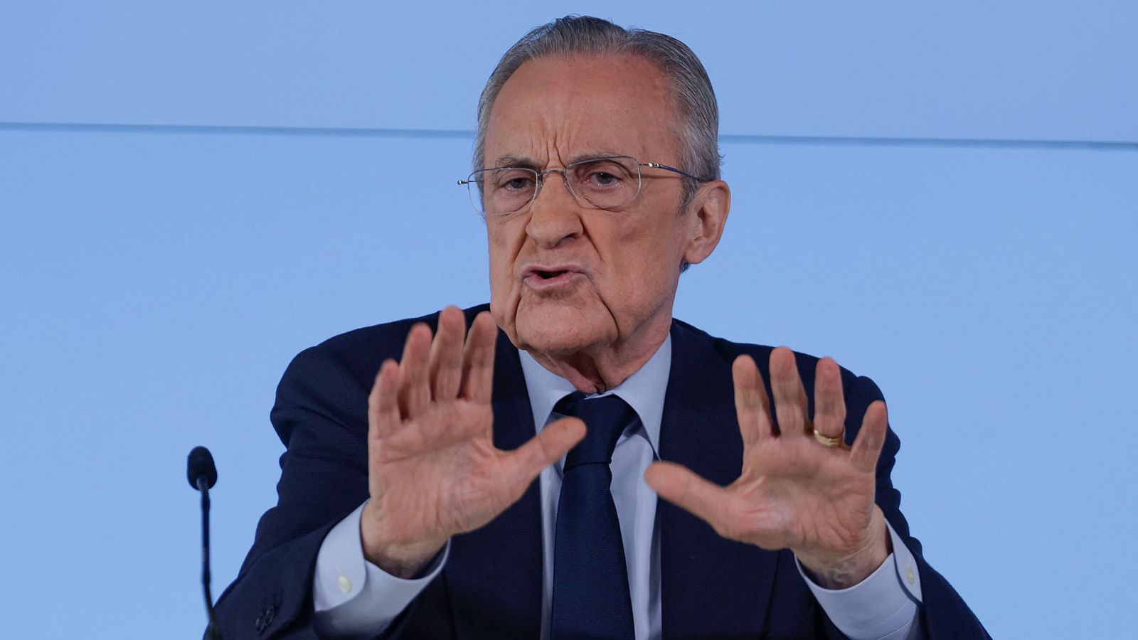 Primer plano de Florentino Pérez, presidente del Real Madrid, en una rueda de prensa. Viste traje azul marino, camisa azul claro y corbata azul oscuro, gesticulando con las manos. Fondo azul claro y micrófono visible.