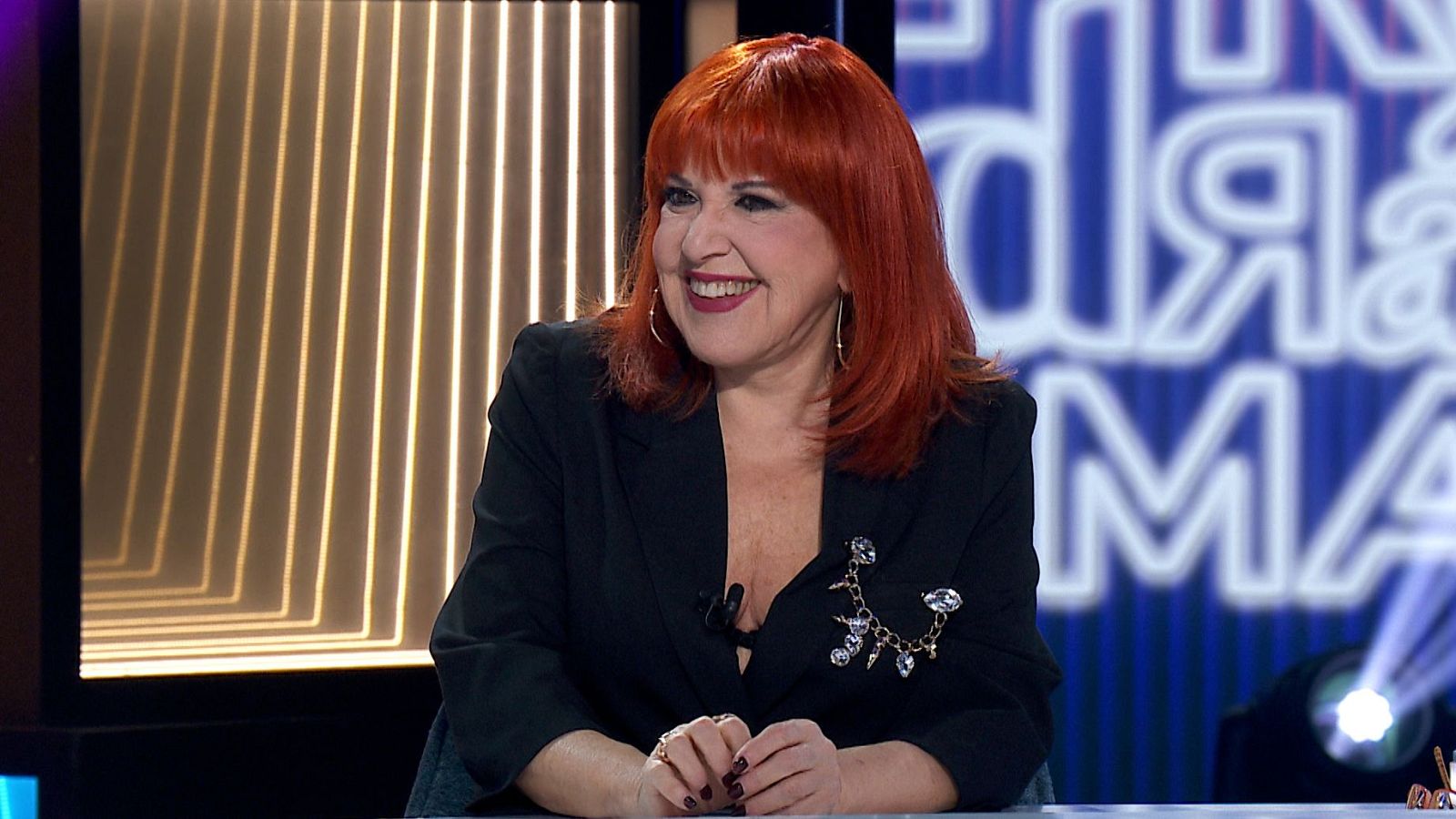 Loles León, con cabello pelirrojo y chaqueta negra, sonríe en un estudio de TV. Lleva un broche dorado y pendientes de aro. Fondo con luces doradas y panel azul con neones "día" y "MA".
