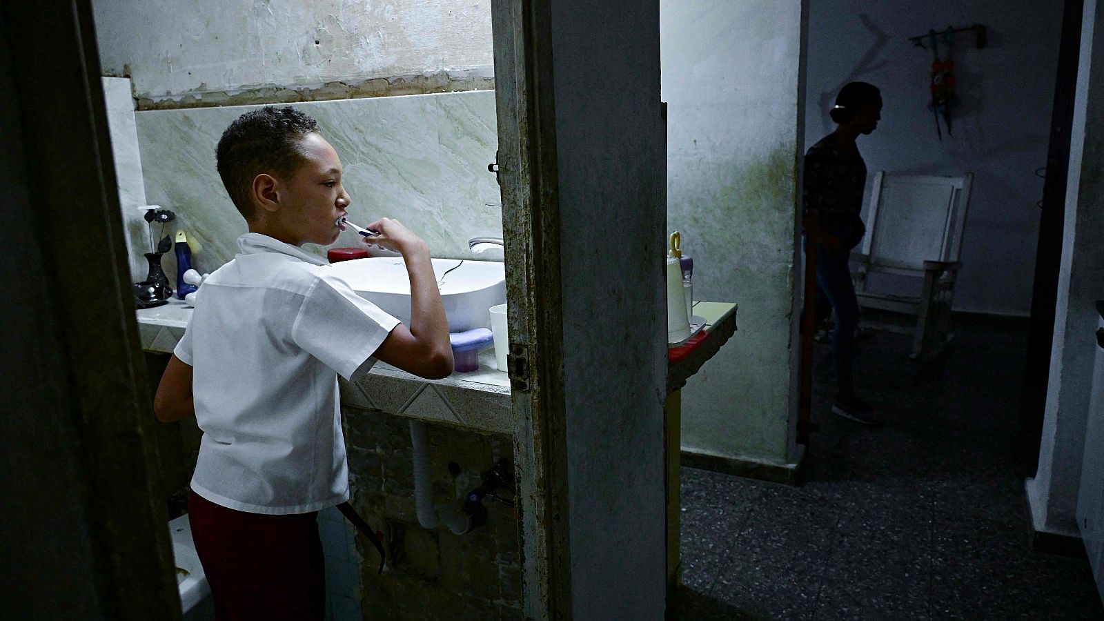 Joven con uniforme escolar se cepilla los dientes en un baño con poca luz. En la habitación contigua, una silueta y una silla mecedora en la oscuridad.