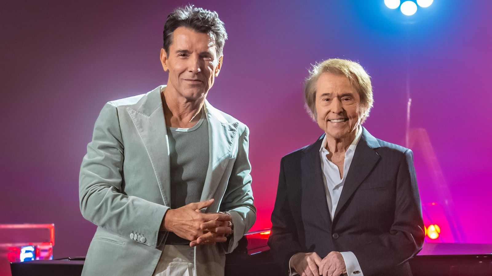 Jesús Vázquez y Raphael, sonriendo, posan en un estudio con fondo degradado morado y rosa. Vázquez viste traje claro, Raphael traje oscuro. Luces de escenario visibles.