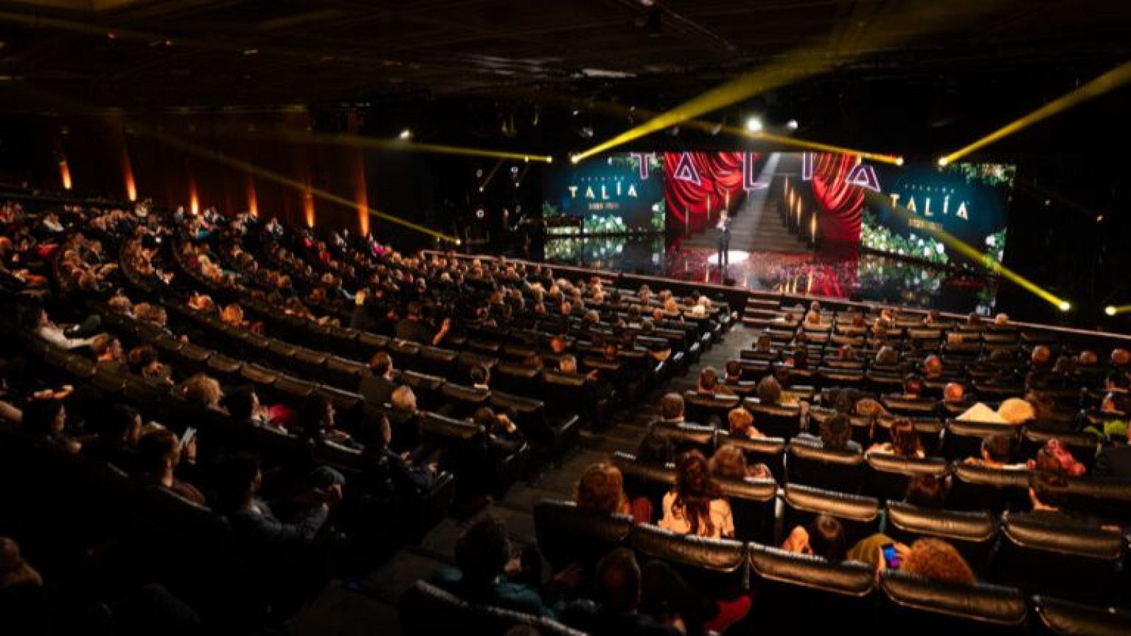Vista de un auditorio con público y escenario iluminado. La pantalla central muestra "TALÍA PREMIOS" con decoración vegetal y cortinas rojas. Una persona se encuentra de pie en el escenario.