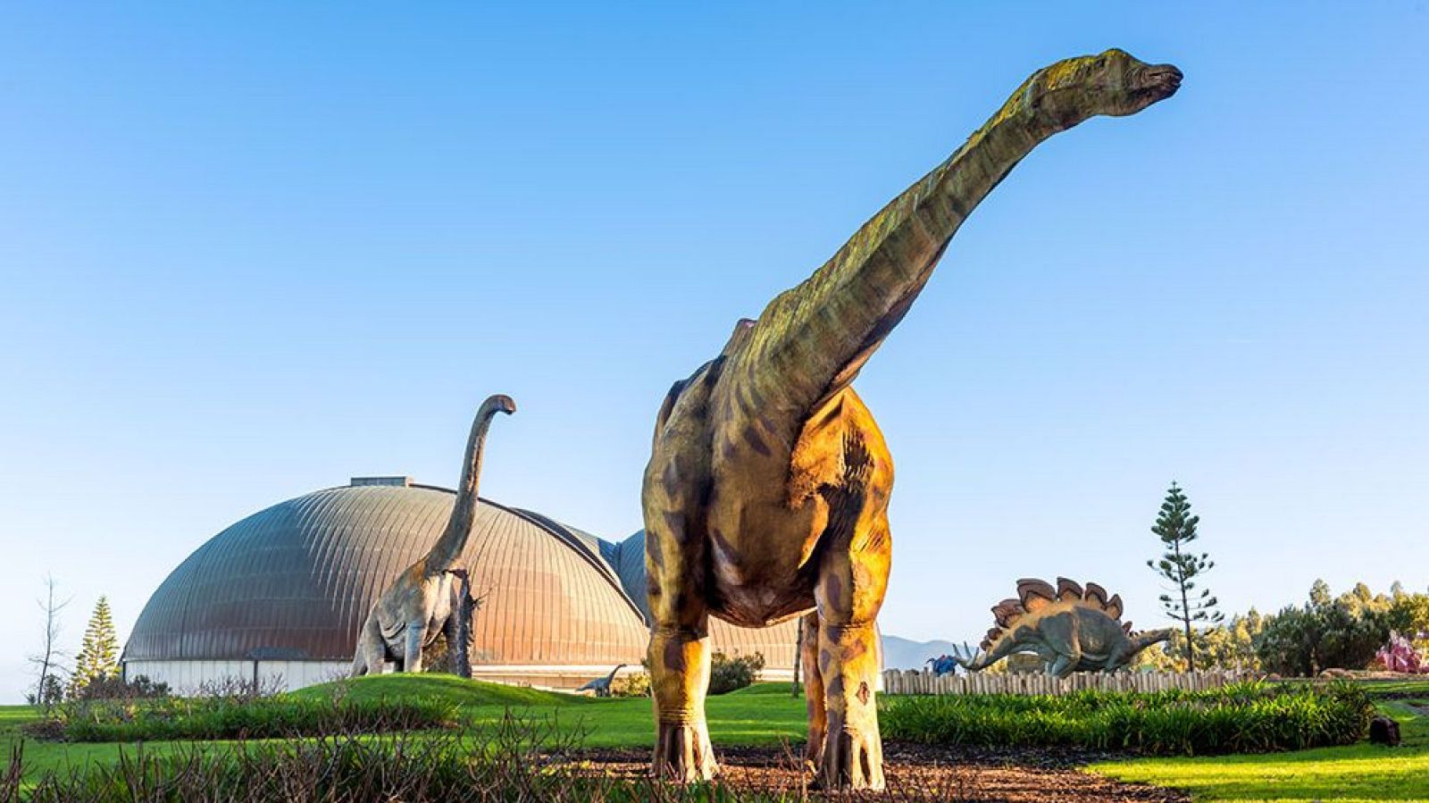 Vista exterior del Museo Jurásico de Asturias con estatuas de dinosaurios, incluyendo un saurópodo y un Stegosaurus, y su icónico edificio abovedado bajo un cielo azul.