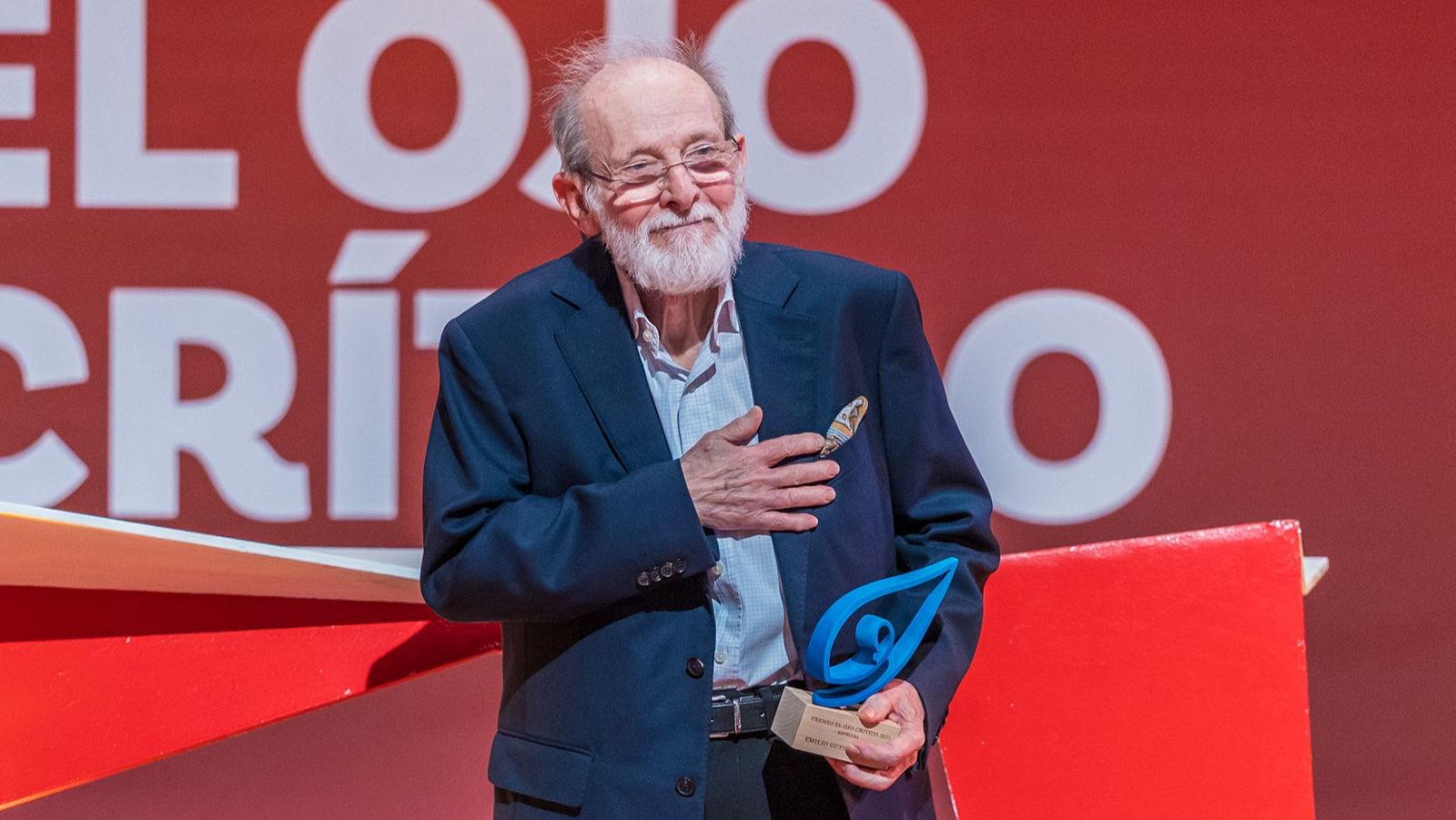Emilio Gutiérrez Caba, actor español, recibe un premio de "EL OJO CRÍTICO". Luce un traje azul marino, barba blanca y gafas, con la mano en el corazón.