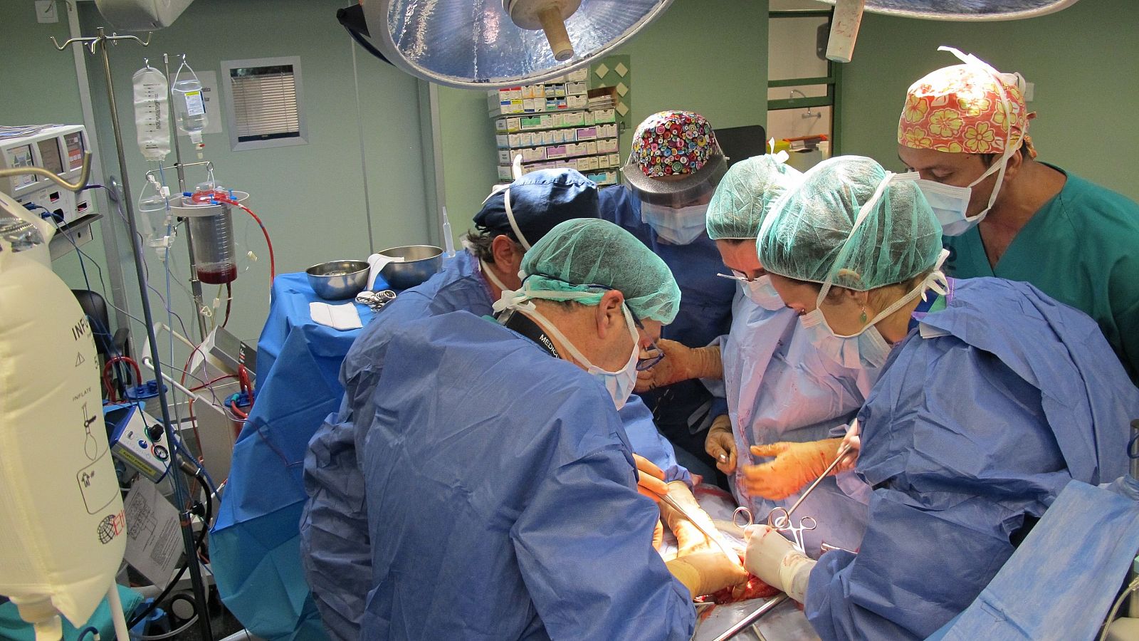 Cirujanos con batas azules, gorros y mascarillas trabajan en un quirófano moderno, rodeando a un paciente. Equipamiento médico y lámparas quirúrgicas iluminan la escena de alta concentración.