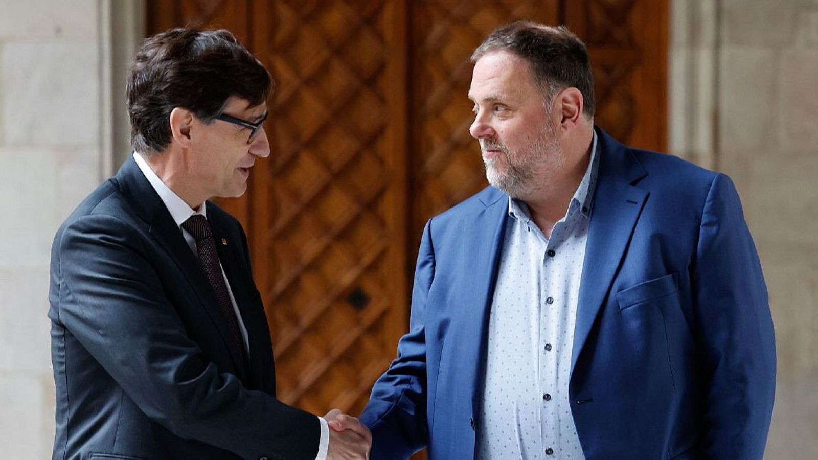 Salvador Illa y Oriol Junqueras se dan la mano en un saludo formal. Illa viste traje oscuro y gafas, Junqueras lleva chaqueta azul y barba. Fondo de madera y piedra.