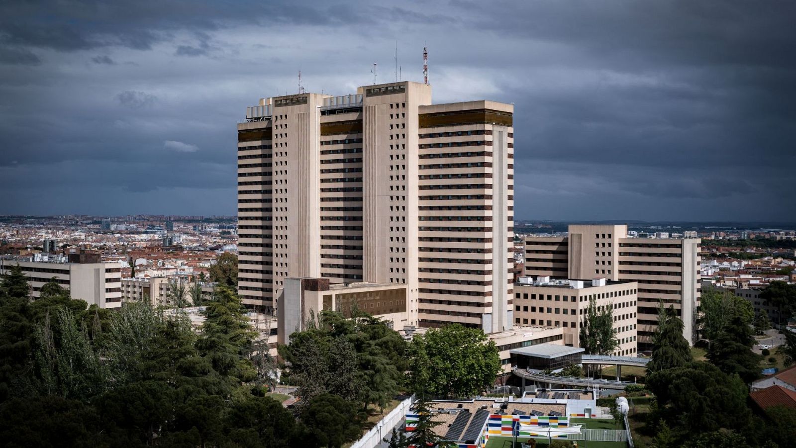 Complejo de edificios del Hospital Gómez Ulla en Madrid, con torres altas y rectangulares, rodeado de vegetación y bajo un cielo nublado.