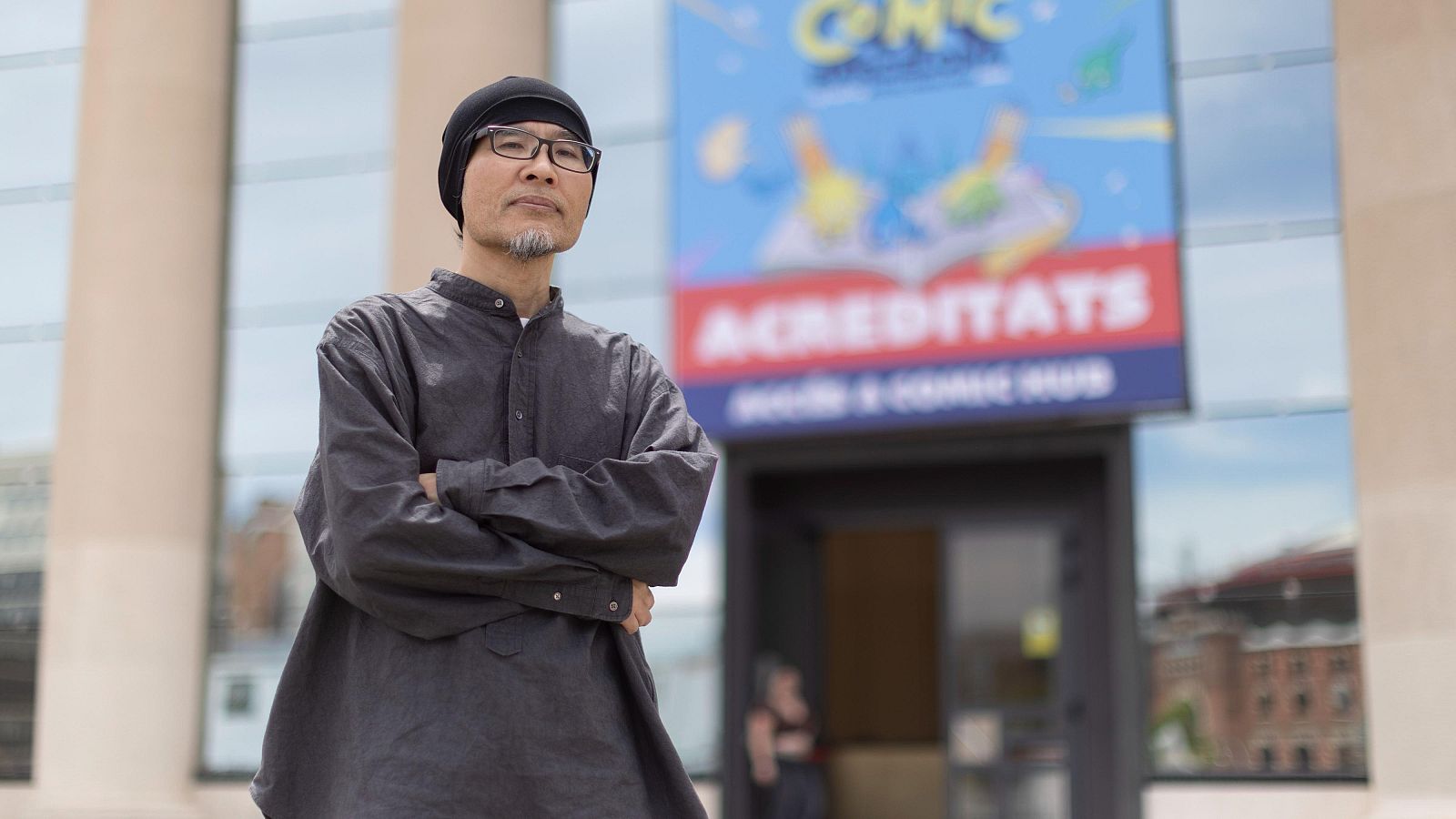 Shitaro Kago, artista de manga, con gorro y gafas, posa frente a un edificio con un cartel que dice "ACREDITATS MANGA & COMIC HUB".