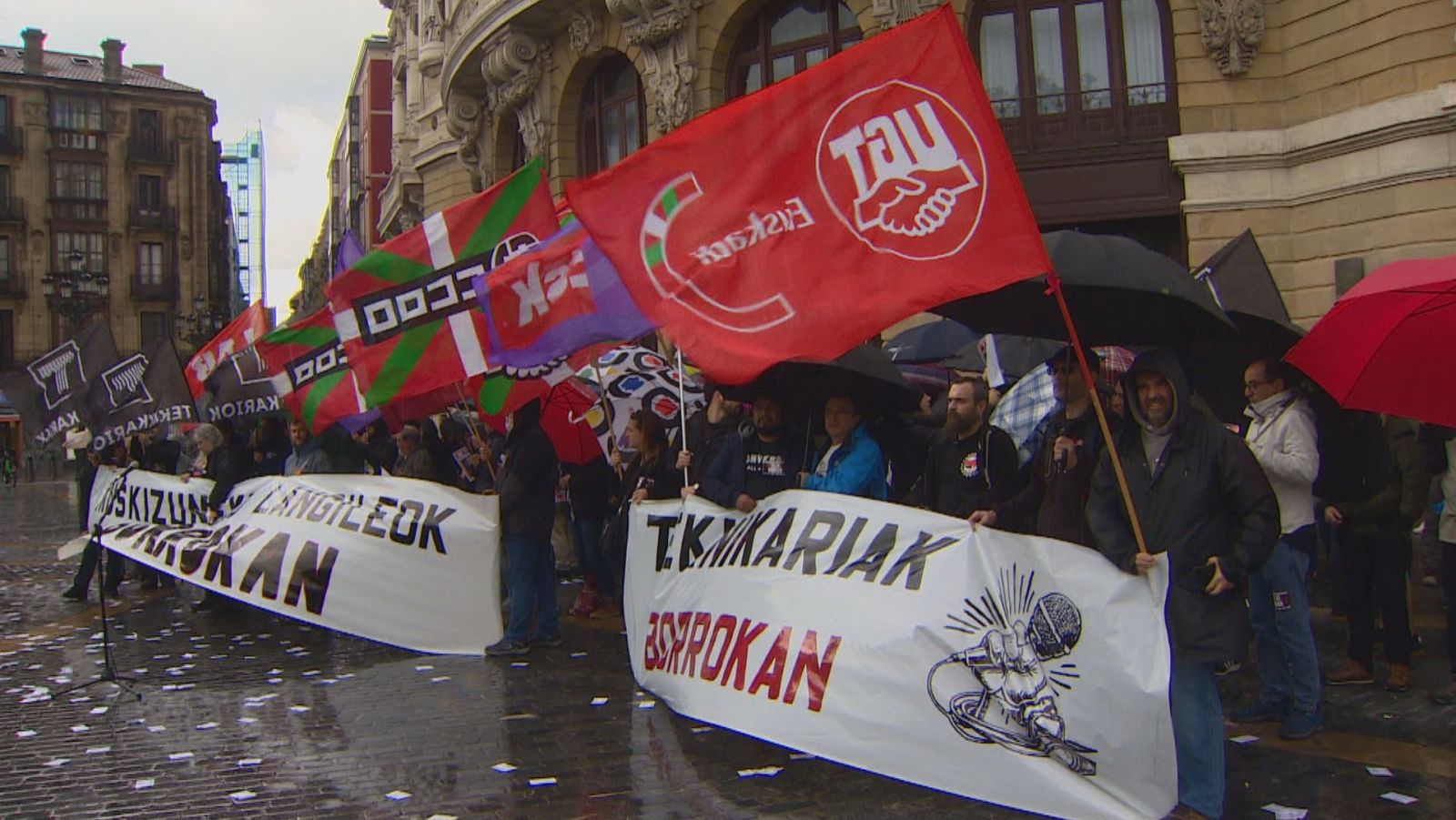 Concentración de trabajadores en una plaza urbana con paraguas y pancartas de sindicatos ELA y LAB, y la Ikurriña. Mensajes en euskera sobre derechos y lucha obrera.