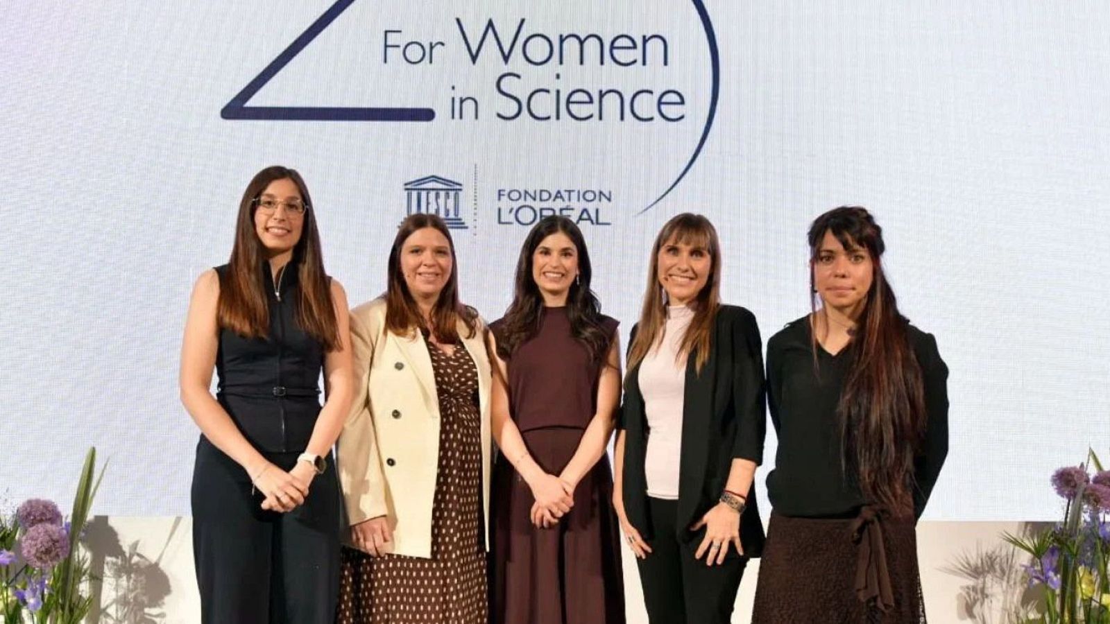 Cinco mujeres científicas, Ylenia Jabalera, Cintia Folgueira, María Puertas-Bartolomé, Ujué Fresán y Sara Mederos, celebran en un evento "For Women in Science" con los logos de UNESCO y L'Oréal.