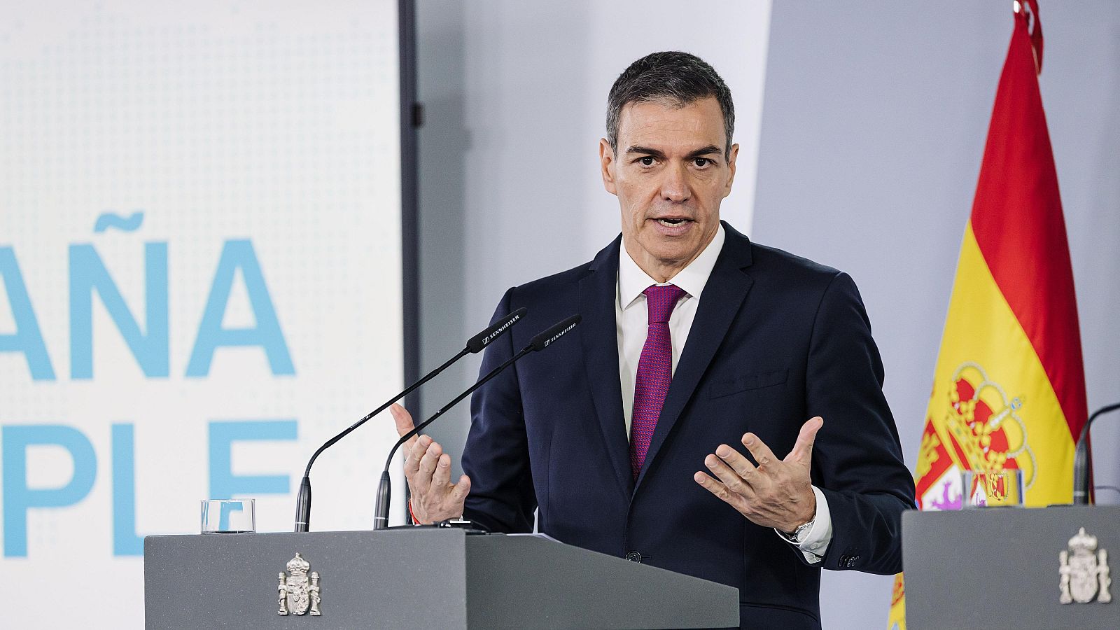 Pedro Sánchez, Presidente del Gobierno, en una comparecencia oficial con la bandera de España y el escudo nacional. Gesticula mientras habla ante micrófonos.