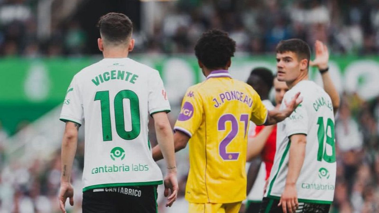 Tres futbolistas en acción: Iñigo Vicente (10) y CANALES (18) con camisetas blancas/verdes, y J. PONCEAU (21) con camiseta amarilla/morada. Se aprecia una interacción entre CANALES y J. PONCEAU.
