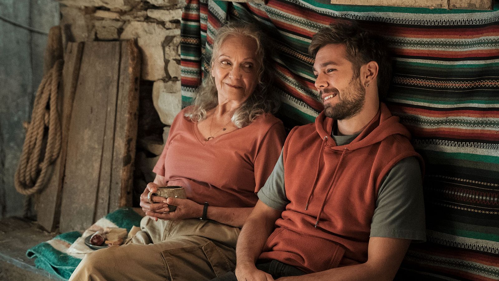 Lola Dueñas y Pol Monen sentados en un ambiente rústico, compartiendo una sonrisa y miradas de conexión. Lola sostiene una taza, mientras Pol luce una sudadera sin mangas.