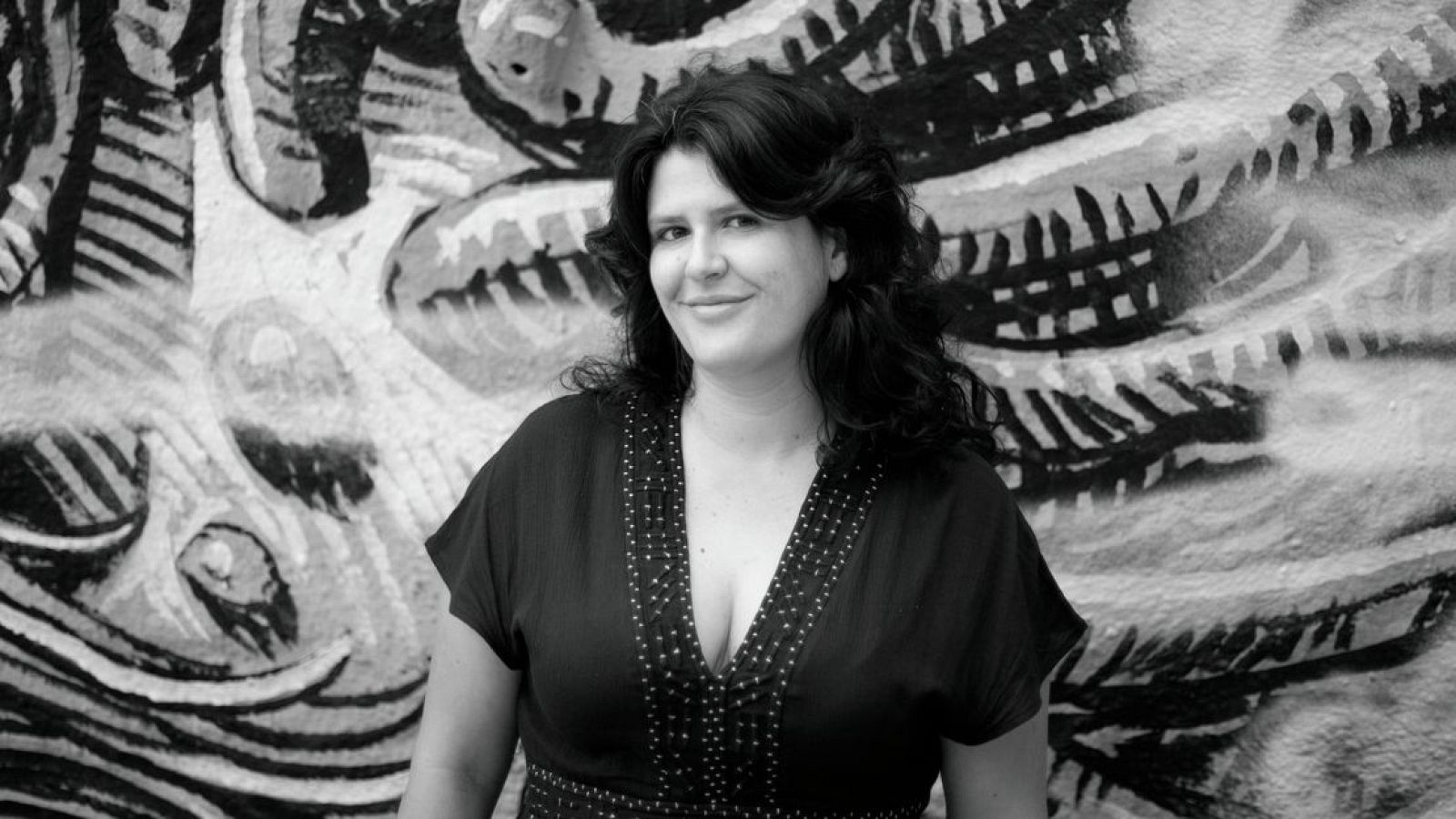 Nuria Núñez Hierro sonríe a la cámara en un retrato en blanco y negro, luciendo una blusa oscura con escote adornado y un fondo de mural abstracto.