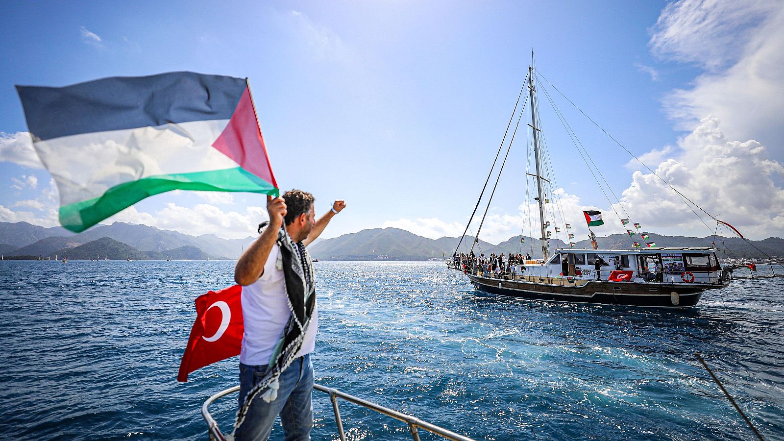 Hombre con bandera de Palestina y pañuelo palestino celebra en un barco. Bandera de Turquía presente. Otro barco con personas y banderas palestinas y turcas al fondo.